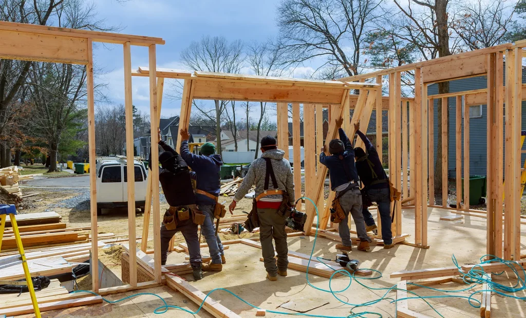Construction workers lifting and positioning a large wooden header frame on a new residential house framing project against a blue sky